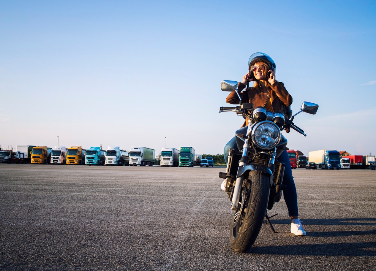 Frau sitzt mit Helm auf einem Motorrad auf einem großen Parkplatz, im Hintergrund stehen Lkw.
