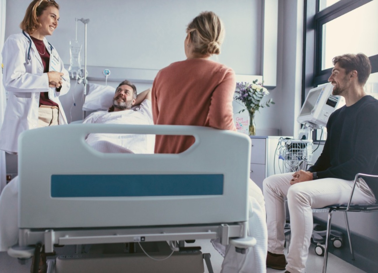 Bühnenbild Ratgeber Krankenhausknigge Ein männlicher Patient liegt in einem Krankenhausbett. Rechts von ihm steht eine weibliche Ärztin, eine Frau sitzt am Fußende des Bettes und ein Mann daneben auf einem Stuhl.