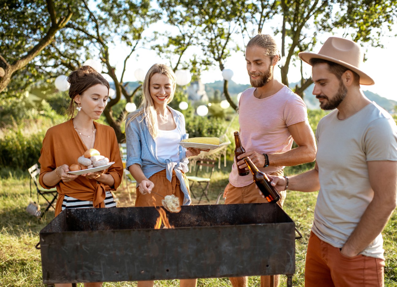 Bühnenbild Ratgeber Sicher Grillen Eine Gruppe von Freunden steht zu viert an einem Holzgrill im Grünen.