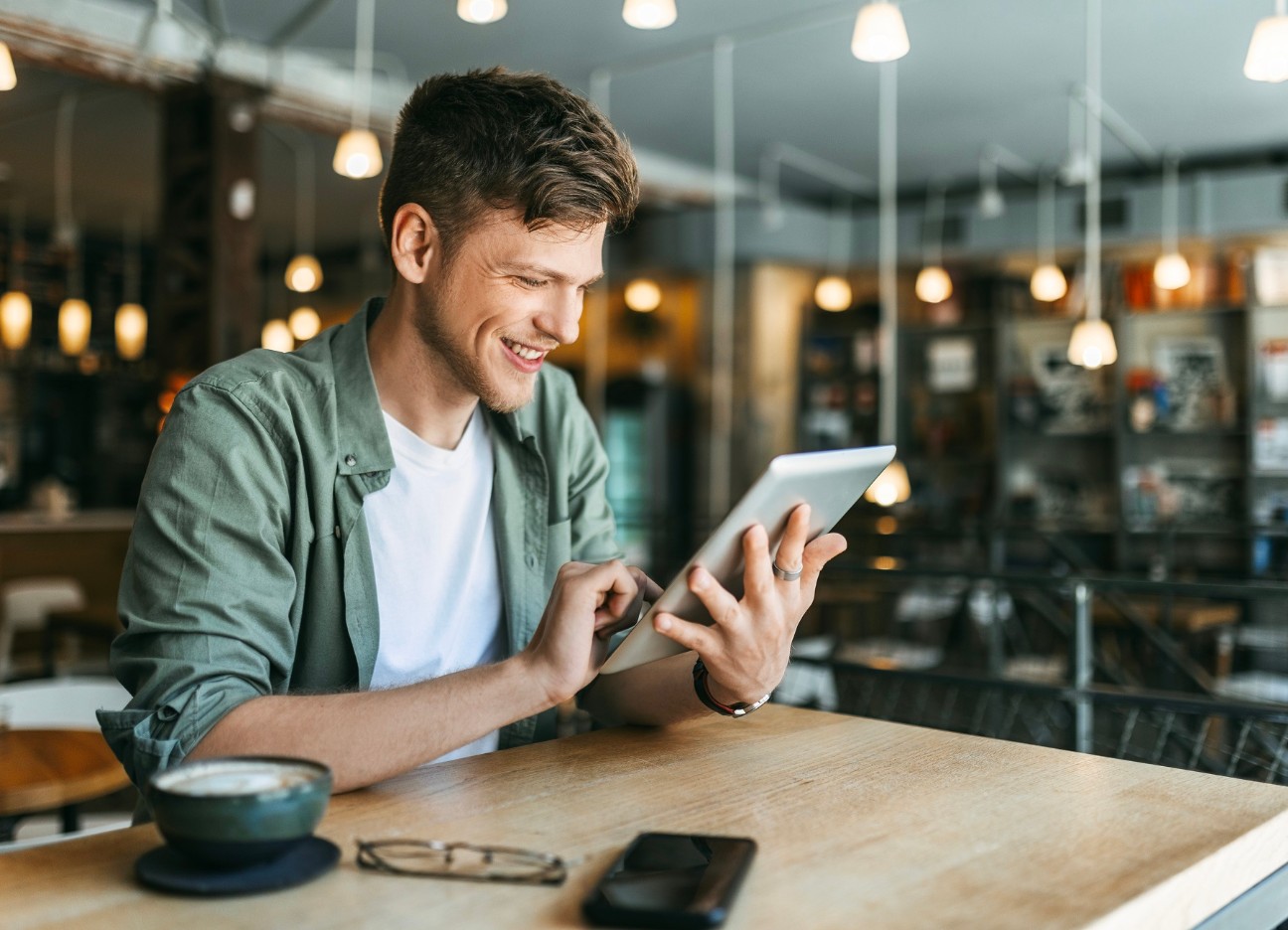 Junger Mann mit grünem Hemd liest in einem Bücher-Cafe auf einem Tablet und lächelt