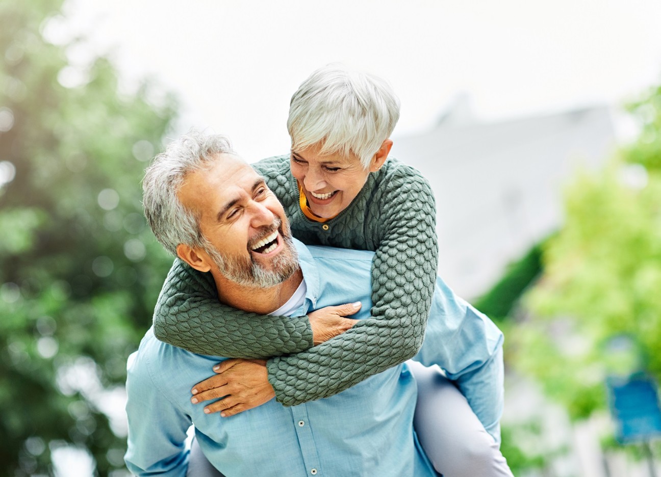Eine ältere Frau sitzt bei einem älteren Mann Huckepack. Beide lachen.