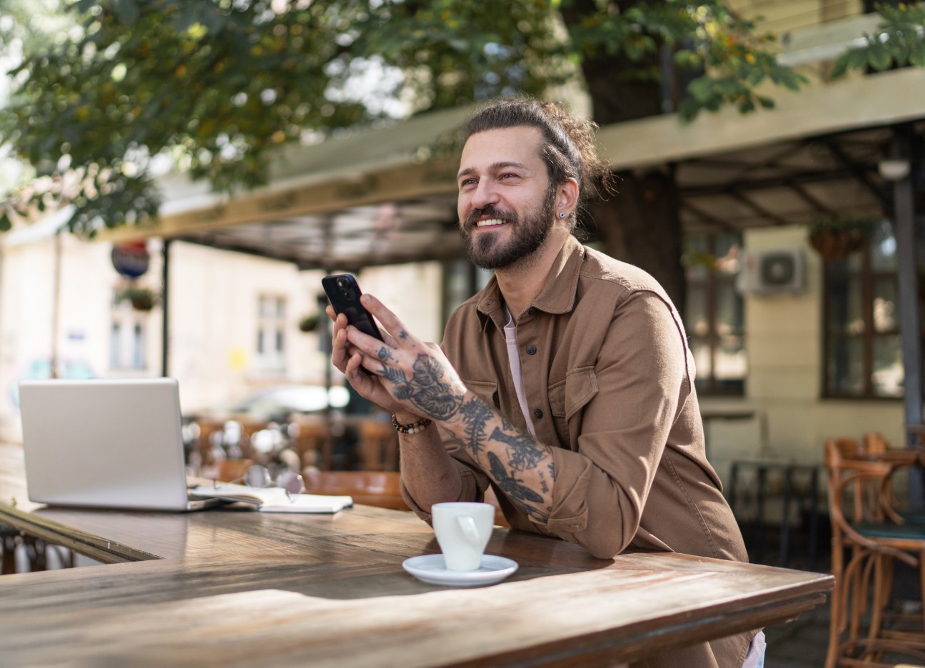 Ein Mann mit Tattoos am Arm sitzt im Außenbereich eines Cafés, hat eine Tasse vor sich stehen und blickt lachend Richtung Straße.