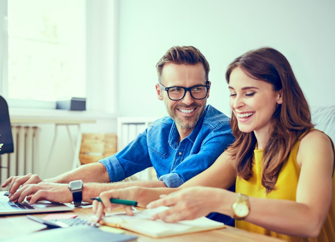 Bühnenbild Ratgeber Investmentsparen Ein Mann und eine Frau sitzen nebeneinander und unterhalten sich lachend, während sie an Unterlagen und am Laptop arbeiten.