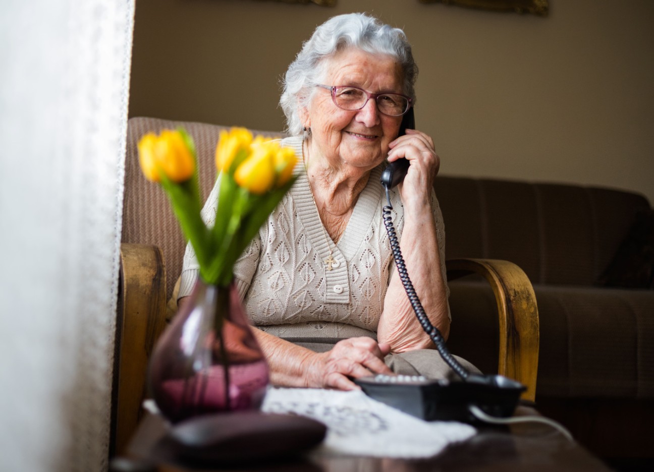 Bühnenbild Ratgeber Trickbetrug Eine ältere Dame sitzt in ihrem Zuhause am Kabeltelefon.