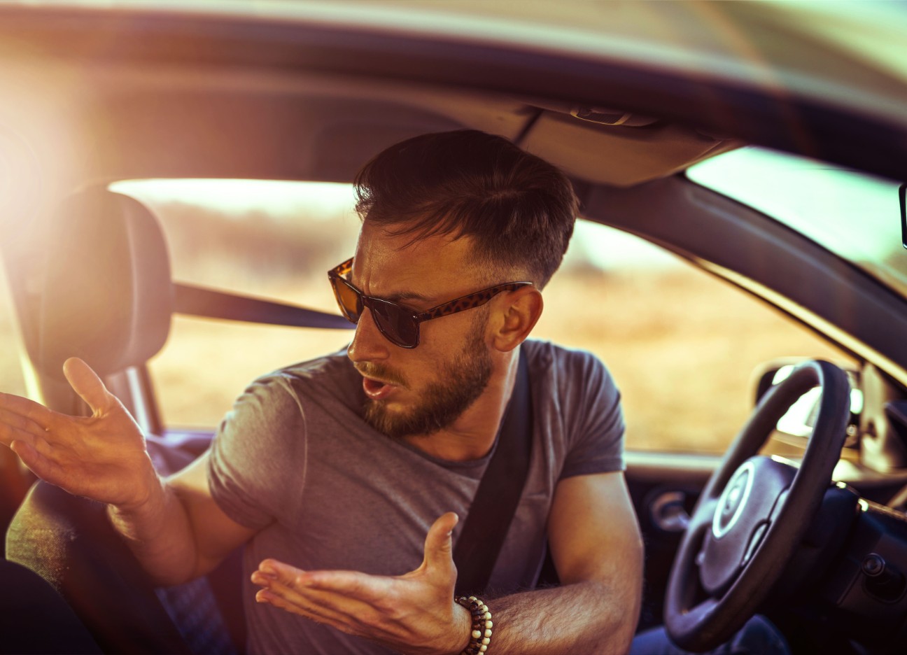 Ein Mann mit Sonnenbrille schaut verwundert und Schultern zuckend um sich im Auto.  