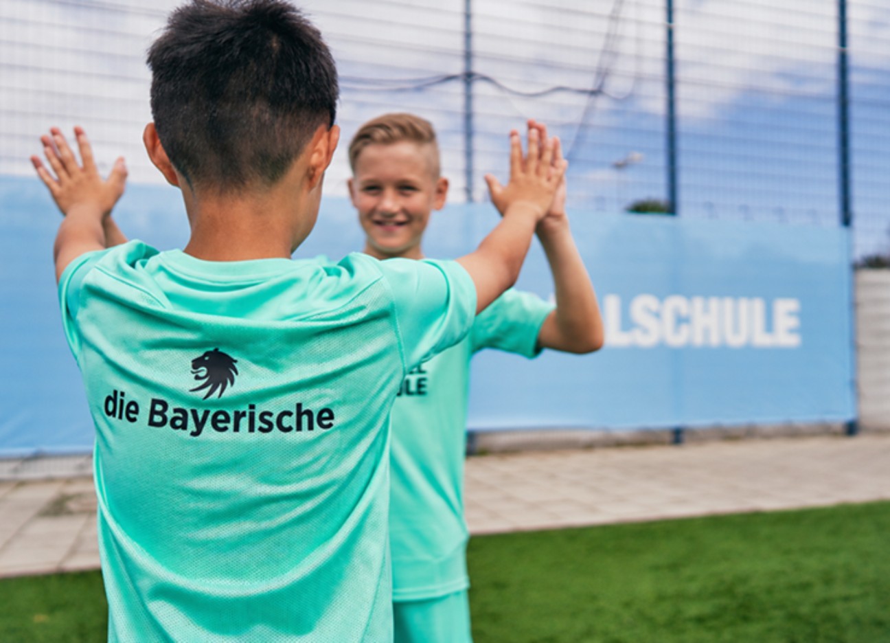 Zwei Kinder in türkisfarbenen Fußballtrikots geben sich auf einem Sportplatz ein High Five. Auf einem Trikot ist das Logo „die Bayerische“ zu sehen.