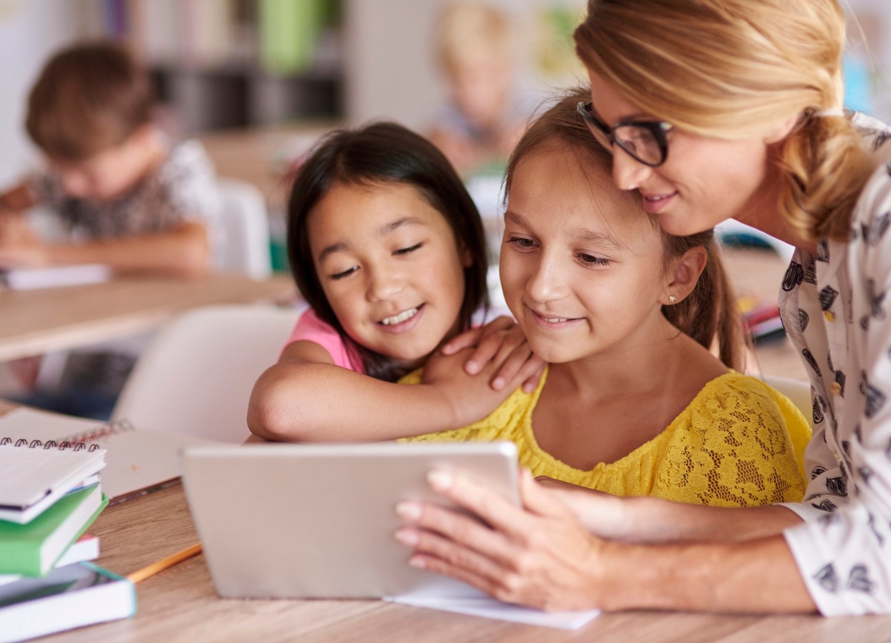 Eine Lehrerin zeigt zwei jungen Schülerinnen etwas auf dem Tablet. Sie sitzen in einem Klassenzimmer.