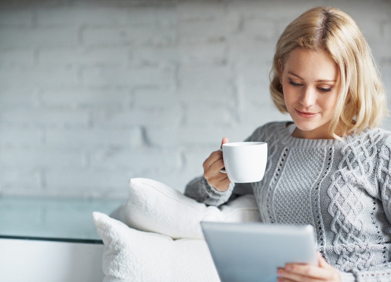 Person sitzt auf einem Sofa, hält eine Tasse und ein Tablet, trägt einen grauen Pullover. Person sitzt auf einem Sofa, hält eine Tasse und ein Tablet, trägt einen grauen Pullover.