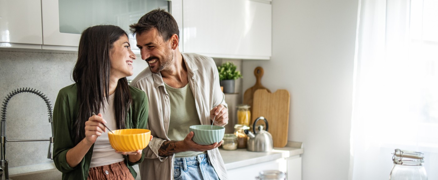 Ein junges Paar steht mit Müslischalen in der Hand steht lachend in der Küche
