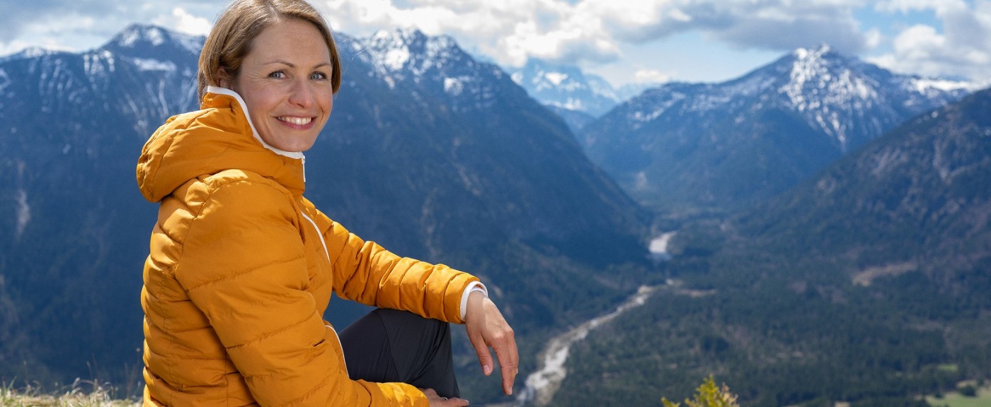 Magdalena Neuner sitzt auf einer Wiese in den Bergen und blickt in die Kamera.