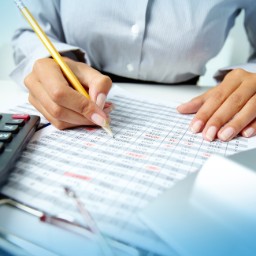 In blau gehaltenes Bild einer großen Kalkulationstabelle auf einem Schreibtisch mit Händen darüber, die einen Stift halten