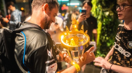 E-Sport-Teilnehmer hält einen goldenen Siegerpokal beim Primehome Cup, während ein Spieler im BIG-Trikot danebensteht.