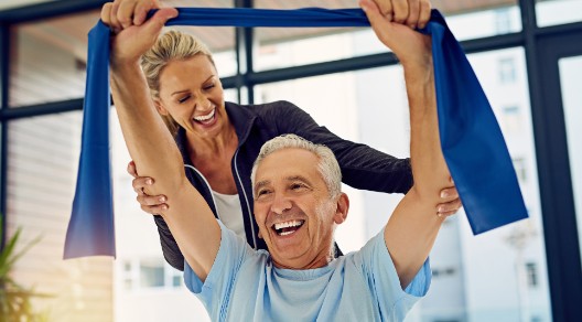 Ein glücklicher, älterer Herr führt mit einem Band eine Physioübung aus, während seine Physiotherapeutin ihn im Hintergrund stützt. 