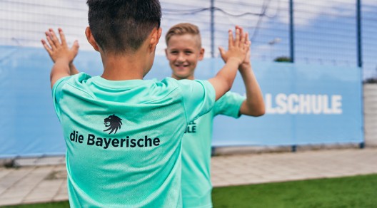Zwei Kinder in türkisfarbenen Fußballtrikots geben sich auf einem Sportplatz ein High Five. Auf einem Trikot ist das Logo „die Bayerische“ zu sehen.