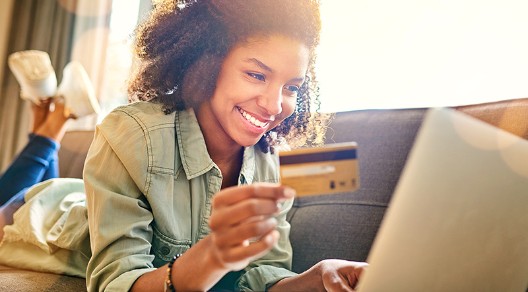 Eine junge Frau liegt auf dem Sofa und tippt lächelnd die Daten ihrer Kreditkarte in ihren Laptop ein.