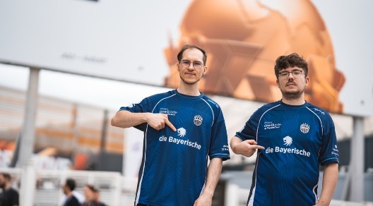 Zwei Männer in blauen E-Sport-Trikots von BIG zeigen auf das Sponsorenlogo „die Bayerische“ auf ihrer Brust; im Hintergrund ist eine große orangefarbene Weltkugel zu sehen.<br/>