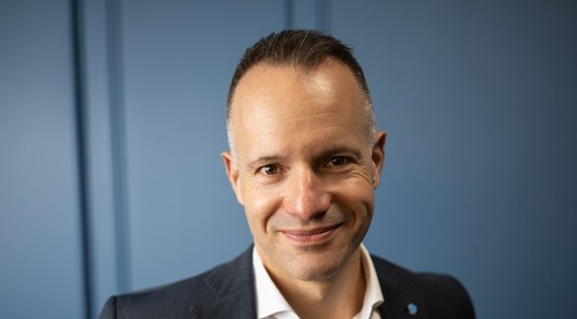 Porträt eines lächelnden Mannes im dunklen Anzug und weißem Hemd vor blauem Hintergrund.