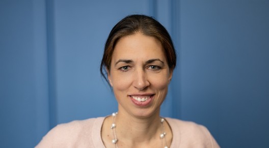 Porträt einer lächelnden Frau mit dunklen Haaren, heller Jacke und Perlenkette vor blauem Hintergrund.