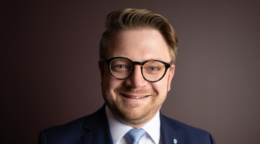 Porträt eines Mannes mit Brille im dunkelblauen Anzug, weißem Hemd und hellblauer Krawatte vor dunklem Hintergrund.