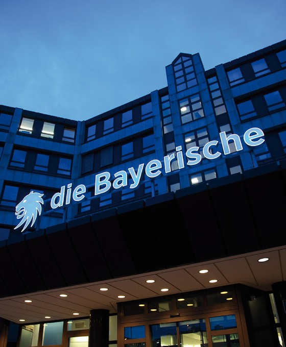 Eingang eines Bürogebäudes mit dem beleuchteten Schriftzug "die Bayerische" bei Dämmerung.