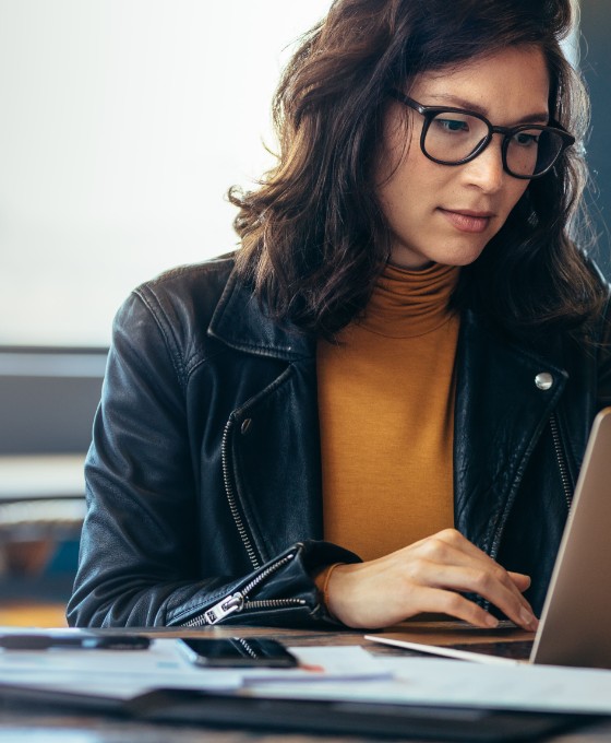 Eine Frau sitzt an einem Tisch und schaut auf ihren Laptop. Neben ihr liegen Dokumente. 