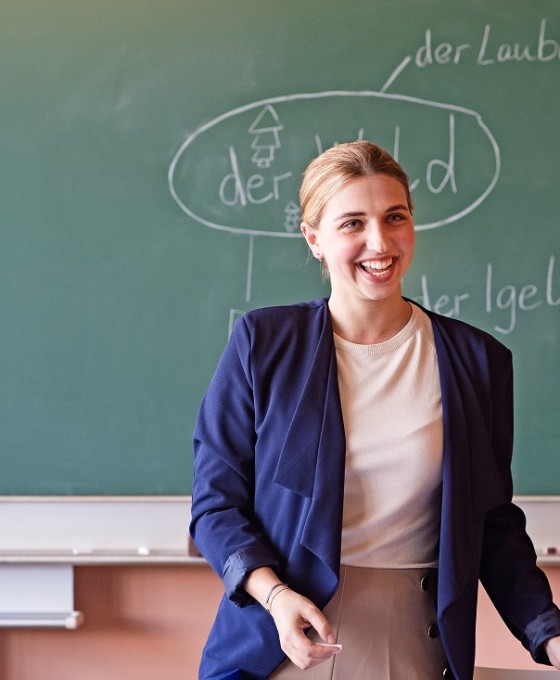 Eine junge Lehrerin steht mit dem Rücken zur Tafel und lächelt