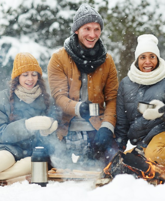 Eine Gruppe von Freunden sitzt in Winterklamotten im Schnee und grillt. 