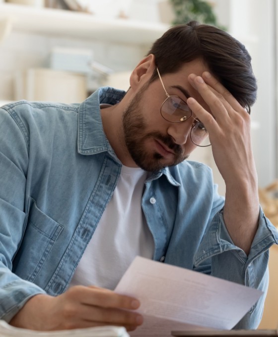Ein Mann sitzt am Laptop und schaut frustriert auf ein kleineres Blatt Papier. 