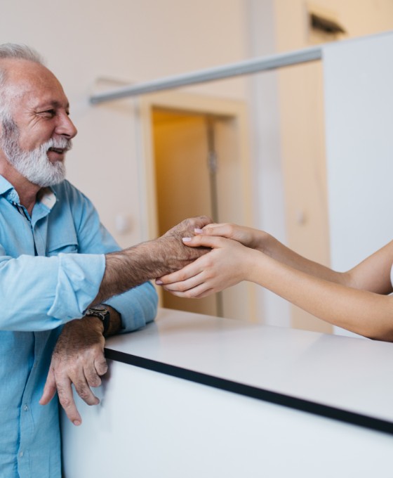 Eine junge Arzthelferin steht hinter der Empfangsrezeption und hält die Hand eines älteren Patienten, der vor ihr am Tresen steht. 