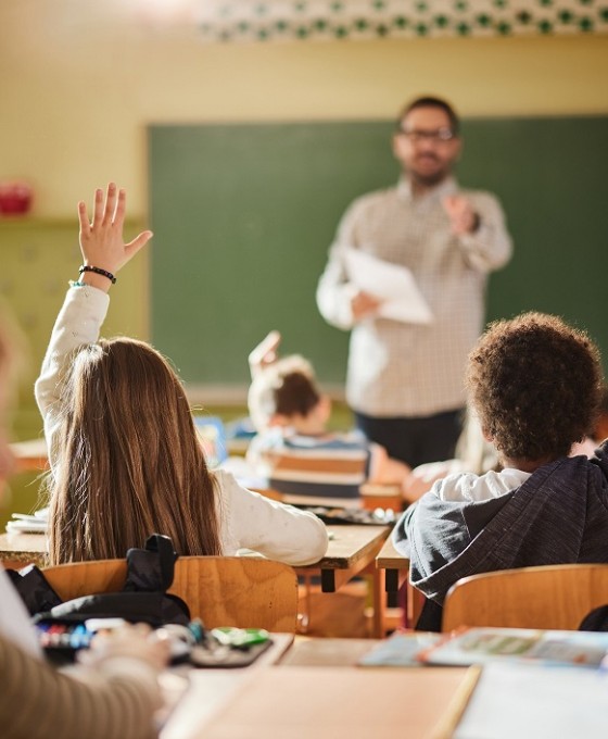Ein Lehrer steht vor einer Klasse von jungen Kindern an der Tafel und fragt etwas ab. Mehrere Kinder melden sich. 