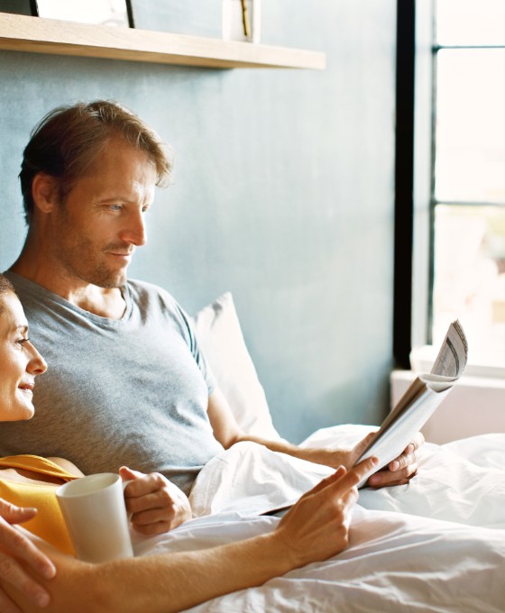 Ein Mann und eine Frau liegen zusammen im Bett und lesen sich eine Zeitung durch. Die Frau hält eine Tasse Kaffee in der Hand. 