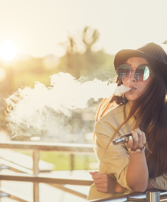 Eine jüngere Frau trägt Hut und Sonnenbrille und raucht eine E-Zigarette.