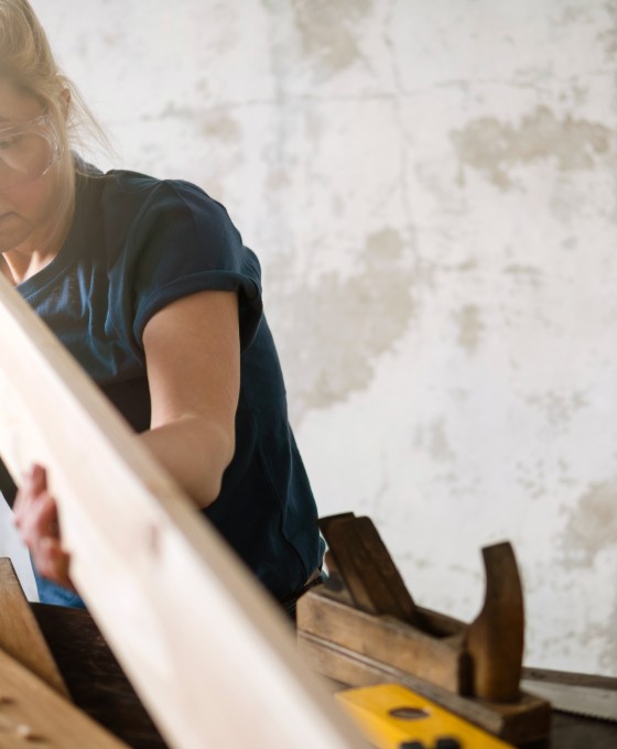 Eine Frau arbeitet an einem länglichen Stück Holz. Sie trägt eine Schutzbrille und neben ihr liegen diverse Werkzeuge. 