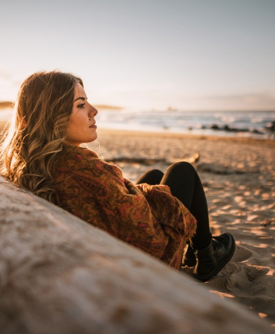 Eine junge Frau sitzt lehnend an einem Holzbalken am Strand. 