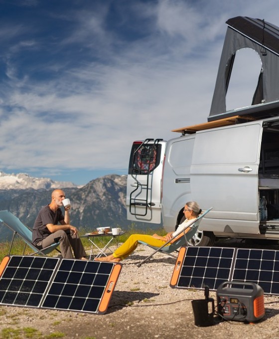 Eine Frau und ein Mann sitzen zusammen in einem Liegestuhl vor ihrem Campervan in den Bergen. Vor ihnen stehen zwei Solarpanels.