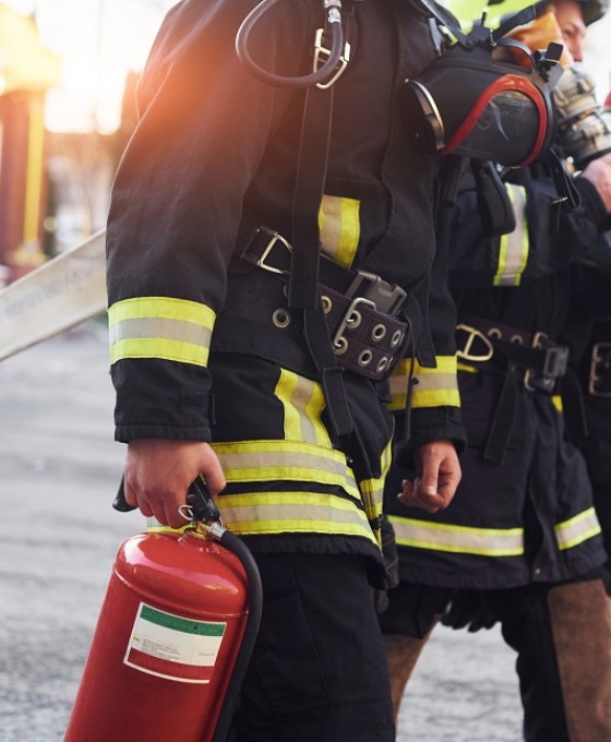 Drei Feuerwehrleute laufen mit Equipment herum. Ihre Uniformen und Equipment stehen im Fokus des Bildes. 