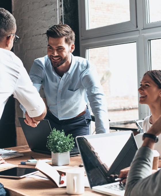 Eine kleinere Gruppe von Leuten sitzen zusammen im Büro und zwei Personen reichen sich die Hände. Sie tragen alle Business-Kleidung. 
