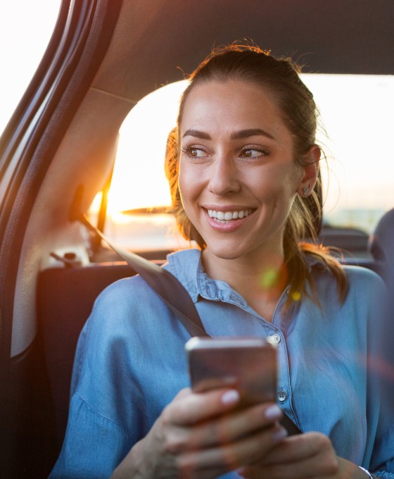 Frau sitzt auf der Rückbank, schaut lachend aus dem Fenster hinaus während sie ein Telefon in der Hand hält.