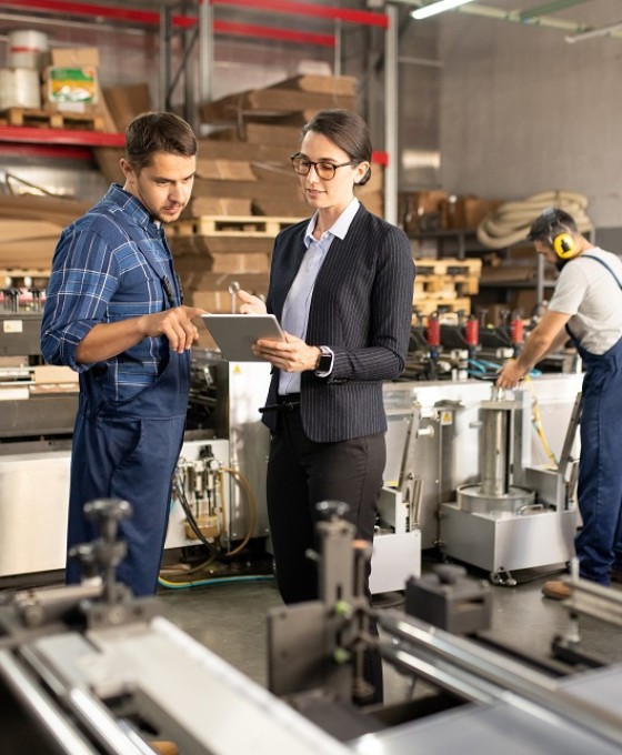 Eine Business-Frau und ein Bauarbeiter sprechen zusammen über ein Dokument, welches die Frau in der Hand hält. Die Umgebung ist eine Fabrik. 