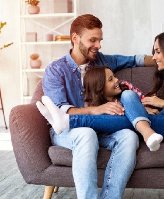 Zwei junge Eltern sitzen lachend mit ihrer Tochter auf dem Sofa.
