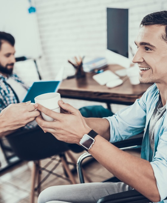 Ein junger Mann sitzt im Rollstuhl und ihm wird eine Tasse Kaffee gereicht. Mit ihm sitzt ein Kollege im Büro. 
