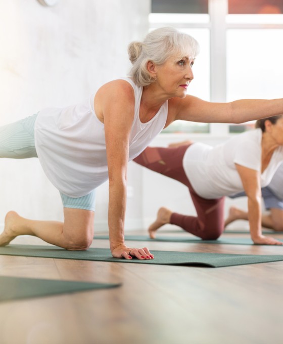 Ältere Frau macht mit anderen eine Balance-Übung auf Yogamatten in einem hellen Raum.