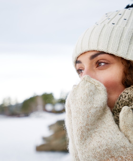 Eine warm eingepackte Frau mit Mütze, Schal und Handschuhen steht draußen in einer verschneiten Landschaft und hält die Hände vor ihr Gesicht, um sich zu wärmen.