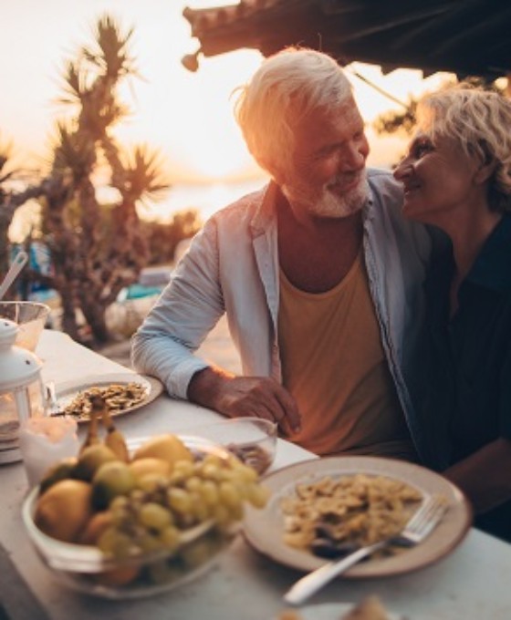 Ein älteres Ehepaar (Rentner) genießen ihr Abendessen unter Palmen im Sonnenuntergang. Sie schauen sich läacheldn an.