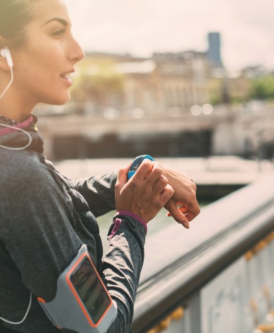 Eine junge Joggerin steht auf einer Brücke und schaut von ihrer Smart Watch hoch. 