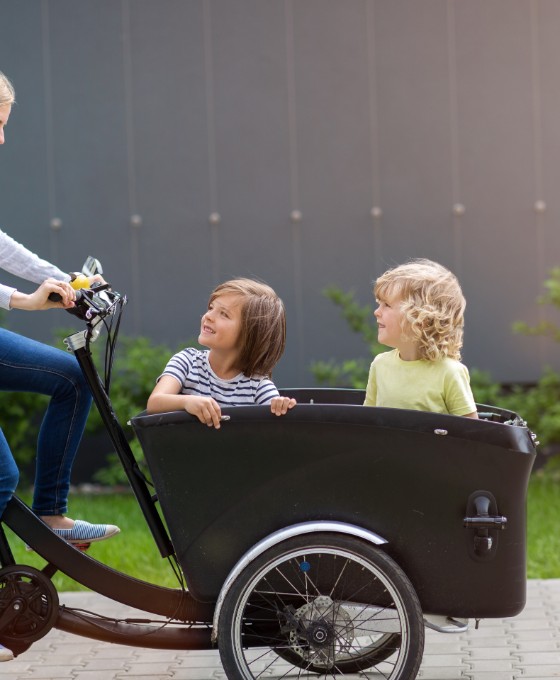 Eine Mutter fährt ein Lastenrad. Vorne im Anhänger sitzen zwei Kinder, die ihre Mutter anschauen. 