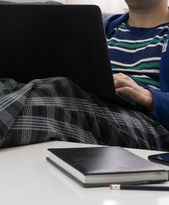 Eine männliche Person (Gesicht nicht zu sehen) liegt auf dem Sofa in Jogginghose und arbeitet am Laptop.