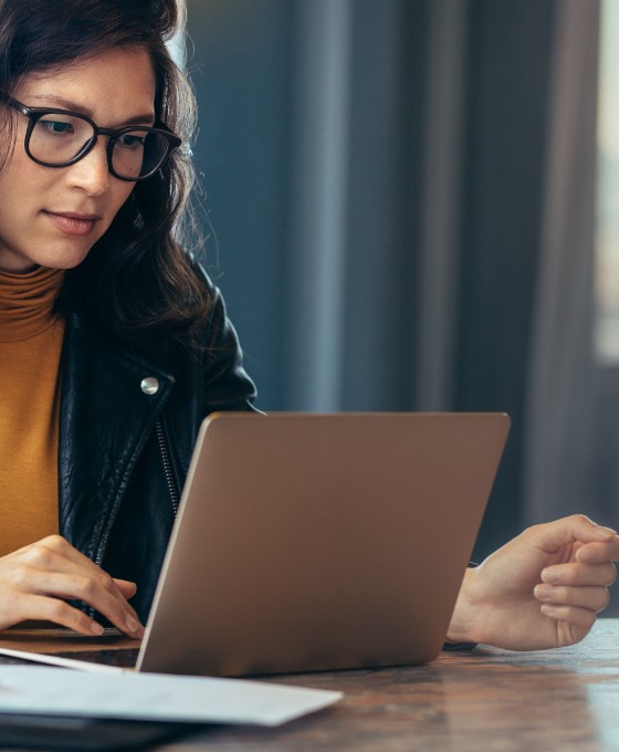 Eine Frau sitzt an einem Tisch und schaut auf ihren Laptop. Neben ihr liegen Dokumente. 