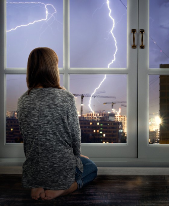 Ein Mädchen sitzt knieend an einem Fenster und schaut die sichtbaren Blitze in der Ferne an. 