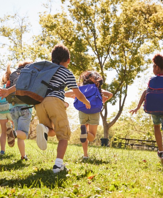 Mehrere Kinder laufen mit ihren Schulrucksäcken über eine Wiese in der Sonne.